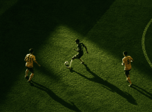 Aerial view of three soccer players on a green field; one in dark kit dribbles the ball while two in yellow defend, with long shadows across the pitch.