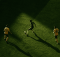 Aerial view of three soccer players on a green field; one in dark kit dribbles the ball while two in yellow defend, with long shadows across the pitch.