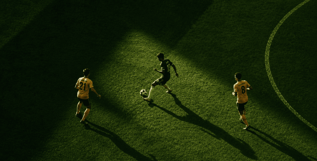 Aerial view of three soccer players on a green field; one in dark kit dribbles the ball while two in yellow defend, with long shadows across the pitch.